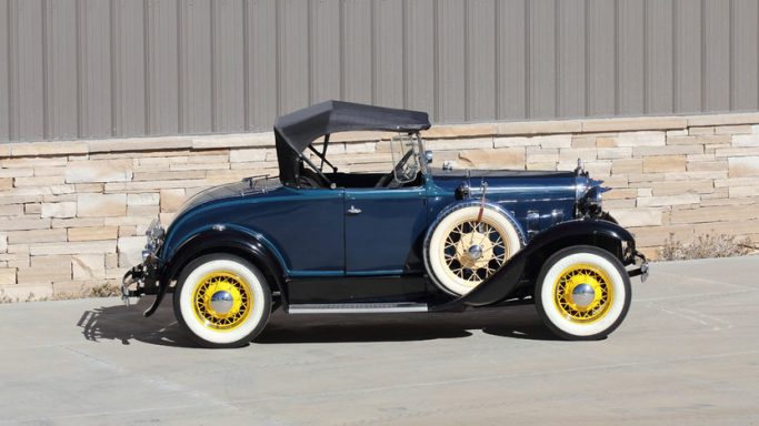 Coche clásico azul con llantas blancas y techo de lona negro, estacionado junto a una pared.